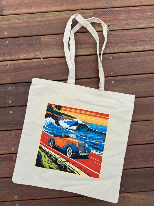 A cream-colored canvas tote bag with a colorful retro graphic of a vintage car on a coastal road.